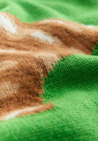 Adītais audums ar zaļām, brūnām un baltām krāsām, kas izceļas ar teksturētu rakstu. Mīksta, pūkaina virsma ar dzīvīgām un blāvām krāsām.