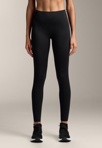 Egy nő magas derekú fekete leggingsben és fekete sportcipőben áll egy sima világos háttér előtt.