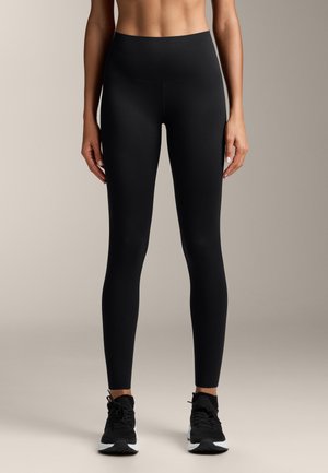 Donna che indossa leggings neri a vita alta e scarpe da ginnastica nere, in piedi dritta contro uno sfondo chiaro e semplice.