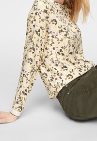 Personne portant une chemise à fleurs jaune, marron et verte à manches longues, assise avec un pantalon vert olive sur une surface gris clair.