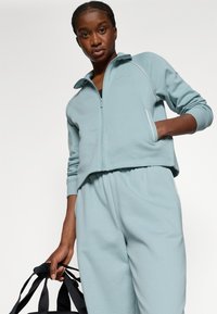 Lyseblå lynlåsfunktion sweatshirt med høj krave og hvid piping, parret med matchende bukser. Stoffet ser blødt og struktureret ud.