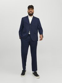 Costum bleumarin cu sacou cu un singur rând de nasturi și pantaloni drepți, asortat cu o cămașă albă și adidași negri. Textură netedă, croială ajustată.