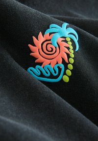 Black fabric features colorful embroidery of a spiral sunflower in pink and orange, with turquoise palm leaves and green accents.