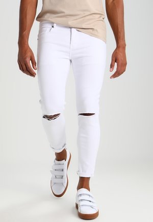 Mand iført slidte hvide skinny jeans, beige top og hvide sneakers med brune såler går foran en ensfarvet baggrund.