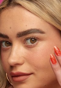 Close-up van een gezicht met lichtbruin huid, prominente wenkbrauwen, bruine ogen en matte lippen, die een hand vasthoudt met lange, glinsterende rode nagels.