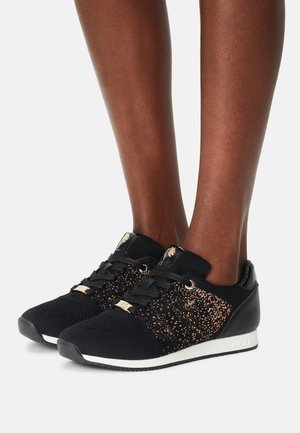 Sneakers in maglia nera con dettagli glitter rosa dorato, lacci neri e suole bianche indossate su gambe dalla pelle scura su sfondo bianco.