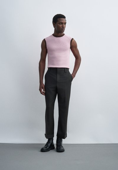 Άνδρας με ροζ αμάνικη μπλούζα με καφέ περίγραμμα, μαύρο παντελόνι με ψηλή μέση και μαύρα δερμάτινα μποτάκια, στέκεται σε ανοιχτό γκρι φόντο.