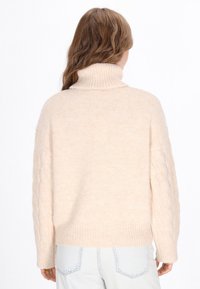 Beiger Oversize-Pullover mit hohem Kragen, versehen mit Zopfmuster-Details auf den Ärmeln und einem gerippten Saum. Weicher, strukturierter Stoff.
