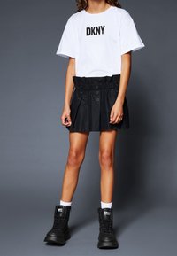 Osoba v bílém tričku DKNY, černé sukni s vysokým pasem a knoflíky, bílých ponožkách a černých šněrovacích botách MM6 na šedém pozadí.