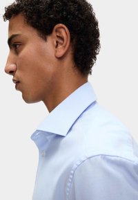 Młody mężczyzna z kręconymi włosami, ubrany w jasnoniebieską koszulę formalną z klasycznym kołnierzykiem, zwrócony w lewo na jednolitym tle.