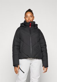 Veste matelassée noire avec un col montant et une capuche, dotée de coutures diagonales et d'une fermeture éclair centrale à l'avant. Accentuée par un tirant de fermeture éclair rouge.
