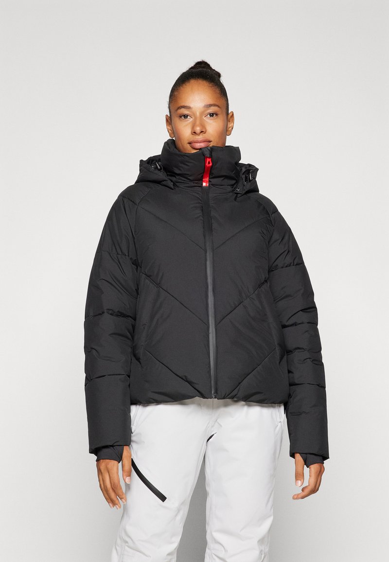 Veste matelassée noire avec un col montant et une capuche, dotée de coutures diagonales et d'une fermeture éclair centrale à l'avant. Accentuée par un tirant de fermeture éclair rouge.
