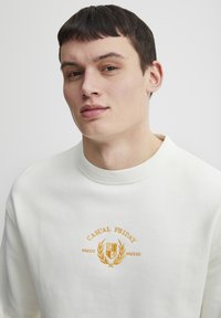 Sudadera blanca con cuello acanalado, logo bordado "CASUAL FRIDAY" en dorado, con acentos de laurel. Textura de tejido suave.