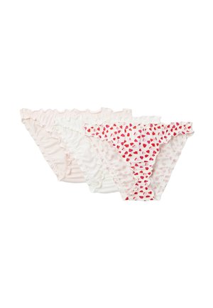 Jeune femme portant un débardeur blanc et des sous-vêtements à motif cœur, debout devant un fond clair uni, regardant vers la gauche.