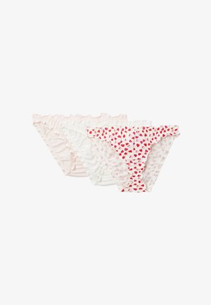 Jeune femme portant un débardeur blanc et des sous-vêtements à motif cœur, debout devant un fond clair uni, regardant vers la gauche.