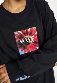 Sort langærmet skjorte med et farverigt blomstertryk og teksten "HUF" placeret mod et blåt kvadrat. Fremstillet i bomuld med rund hals.