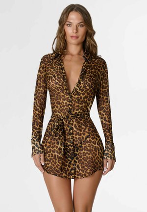 Femme portant une mini-robe à manches longues boutonnée avec un imprimé léopard et une ceinture nouée à la taille, debout devant un fond uni.