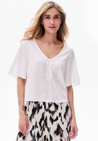 Biała bluzka z krótkim rękawem, dekoltem w serek i dwoma kokardkami z przodu. Lekki materiał i luźny krój. Widać wzorzystą spódnicę poniżej.