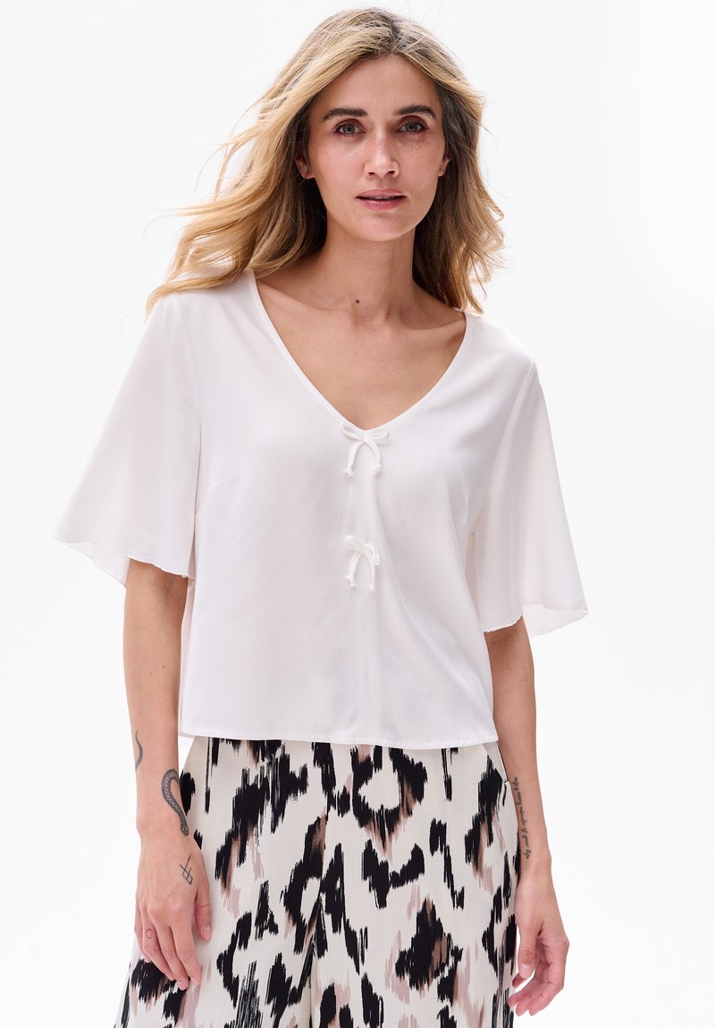 Biała bluzka z krótkim rękawem, dekoltem w serek i dwoma kokardkami z przodu. Lekki materiał i luźny krój. Widać wzorzystą spódnicę poniżej.