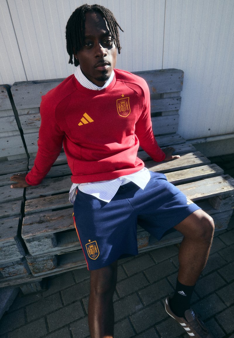 Crvena dukserica s žutim Adidas logotipom i grbom Španjolske, uparena s mornarsko plavim kratkim hlačama s žutim detaljima i logotipom. Prugasta majica ispod.