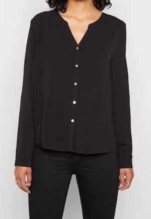 Persoon draagt een zwarte blouse met lange mouwen, knopen en een V-hals, en zwarte broek, staand tegen een effen lichte achtergrond.