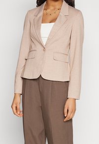 Blazer beige en tissu texturé, présentant une coupe ajustée, des revers à cran, une fermeture à un bouton et deux poches avant.