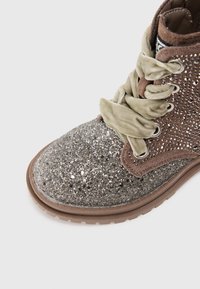Stivale con fibbia, punta argentata e grigia luccicante, parte superiore in suede marrone e lacci verde chiaro. Presenta dettagli metallici sulle asole.