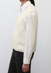 Cremfarbener, gestrickter ärmelloser Pullover mit V-Ausschnitt, kombiniert mit einem weißen Langarmhemd. Dunkle Hosen runden das Outfit ab.