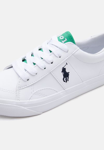 Λευκό αθλητικό παπούτσι χαμηλού προφίλ με λευκά κορδόνια, πράσινη γλώσσα και μαύρο λογότυπο παίκτη πόλο στο πλάι, σε ουδέτερο φόντο.