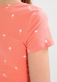 Camisa de manga corta color coral con un patrón de pequeños flamencos blancos, llevada por una persona de piel clara y cabello castaño.