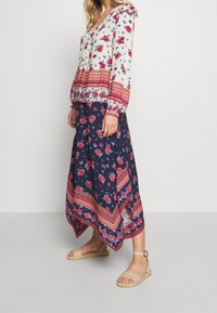 Blouse fleurie avec poignets à volants, fond crème, motif floral rouge et bleu. Jupe longue bleu marine avec imprimé floral et ourlet à motifs, assortie à des sandales beiges.