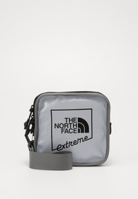 Borsa a tracolla grigia in nylon con chiusura a zip, dotata di un logo nero sulla parte anteriore e di una tracolla grigia regolabile.