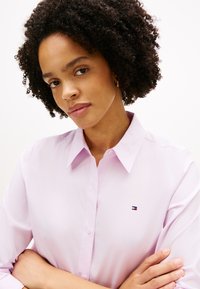 Νεαρή γυναίκα με σγουρά μαλλιά που φοράει ένα ανοιχτόροζ πουκάμισο με κουμπιά και μικρό λογότυπο στο στήθος, με σταυρωμένα τα χέρια, μπροστά σε απλό φόντο.