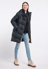 Gepolsteter, dunkelgrauer Mantel mit Kapuze, Reißverschluss vorne und Seitentaschen, ausgestattet mit einem gemusterten Innenfutter und einem orangefarbenen Etikett. Kombiniert mit hellen Jeans und beigen Stiefeln.