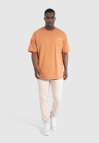 Kurzärmliges, rundhalsiges T-Shirt aus rostfarbenem Baumwollstoff mit weißem Logo, kombiniert mit hellbeigen Sweatpants und weißen Turnschuhen.