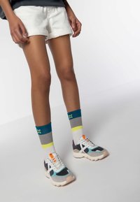 Sneaker colorati realizzati in materiali a rete e camoscio, con una suola spessa, abbinati a calzini a coste in tonalità teal, grigio e con accenti gialli.