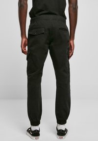 Uomo che indossa pantaloni jogger neri con tasche cargo e polsini elasticizzati, abbinati a calze bianche e sneaker nere, in piedi davanti a uno sfondo neutro.