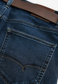 Jeans din denim de un albastru închis, cu un buzunar pe spate ce are detalii de cusătură subtile. O curea din piele maro se află pe talie.