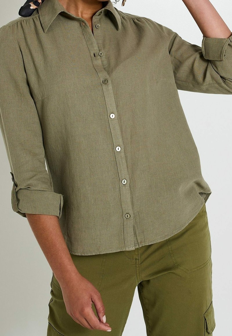 Personne portant une chemise boutonnée vert olive avec les manches retroussées et un pantalon cargo vert olive assorti sur un fond uni.