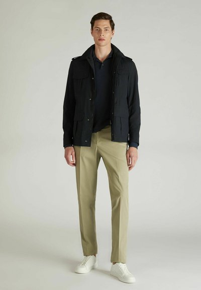 Veste légère noire avec plusieurs poches, associée à un polo sombre et un chino beige, complétée par des baskets blanches. Texture lisse.