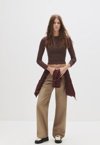 Braunes langärmeliges cropped Top mit rundem Halsausschnitt, beige weite Hosen und schwarze Schuhe mit weißen Akzenten. Das Model hält eine Jacke, die am Hüftbereich gebunden ist.