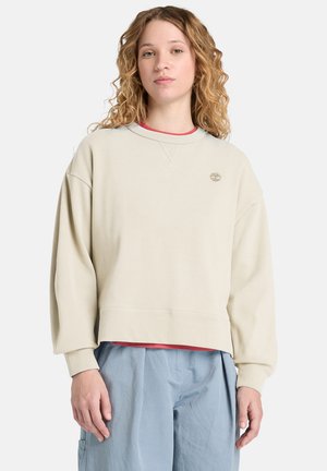 Młoda kobieta o kręconych włosach, ubrana w beżową bluzę z logo i jasnoniebieskie spodnie, stojąca na białym tle.