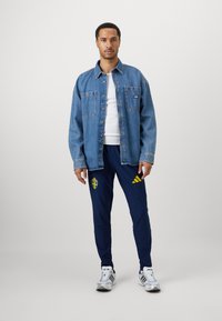 Homem de pé, vestindo uma jaqueta de ganga sobre uma camisa branca, calças azul-marinho com logótipos amarelos e ténis brancos, contra um fundo simples.