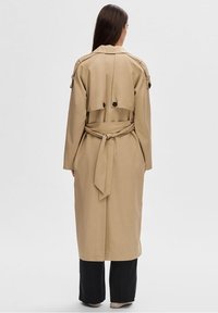 Kvinna med långt brunt hår som bär en beige bältes trench coat och svarta byxor, stående vänd bortåt mot en neutral ljus bakgrund.