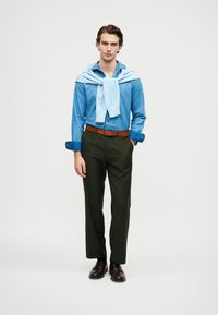 Maglione azzurro chiaro annodato su una camicia blu a bottoni, pantaloni verdi e cintura marrone. Mocassini marroni scuri completano l'outfit.