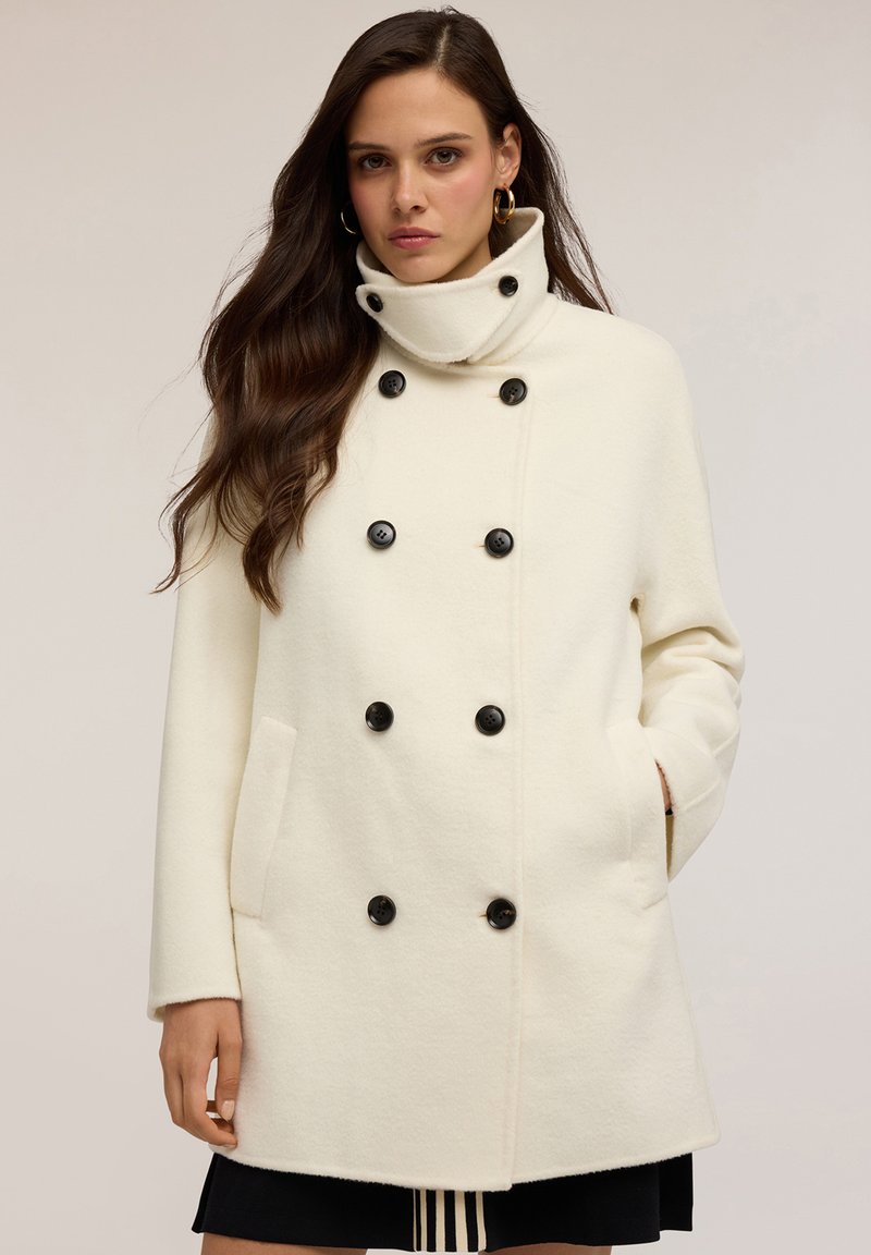 Cappotto doppiopetto color crema realizzato in materiale morbido, con bottoni neri, colletto alto e tasche frontali.