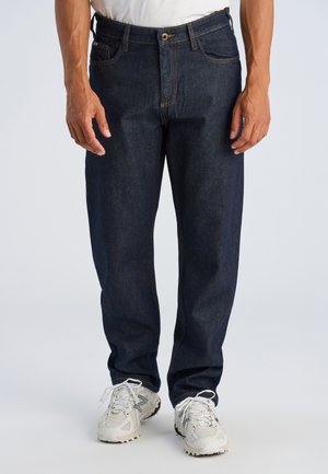 Man draagt donkerblauwe jeans en witte sneakers, staand met ontspannen handen langs zijn zij op een effen achtergrond.