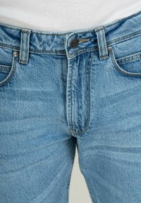 Pantalones vaqueros de denim azul claro con un diseño clásico de cinco bolsillos, costuras visibles, cierres de botón metálico y una textura ligeramente deslavada.