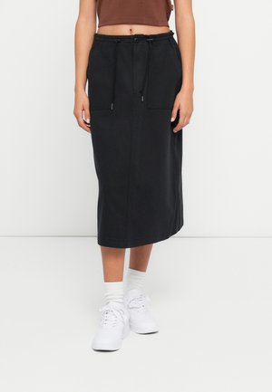 Kvinde iført en sort midi-nederdel med forlommer, en brun cropped top, hvide sokker og hvide sneakers stående på en ensfarvet hvid gulv.