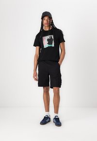 T-shirt graphique noir avec un imprimé coloré, associé à un short cargo noir. Porté avec une casquette noire et des chaussures de sport bleues.